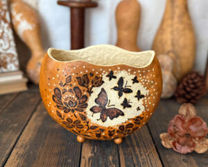 A butterfly themed gourd bowl sits on a distressed wood table. The rim is carved in a wavy shape, the front has butterfly art burned into it